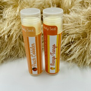 May include: Two lip balm tubes with white caps and orange labels. The labels have the text "Society Seal" and the names of the flavors, "Peach Cobbler" and "Mango Tango".