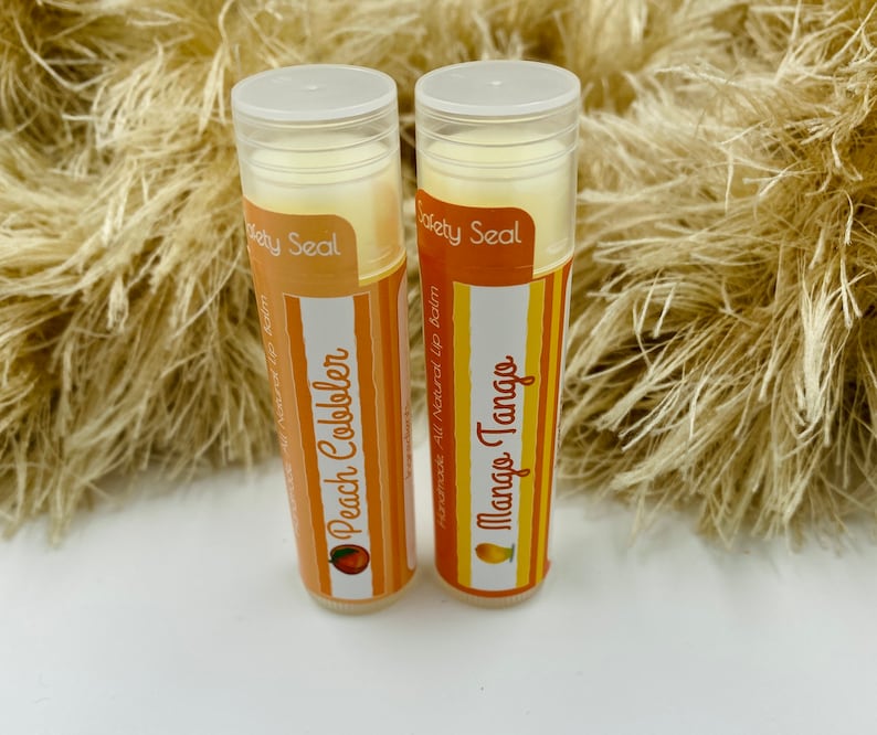 May include: Two lip balm tubes with white caps and orange labels. The labels have the text "Society Seal" and the names of the flavors, "Peach Cobbler" and "Mango Tango".