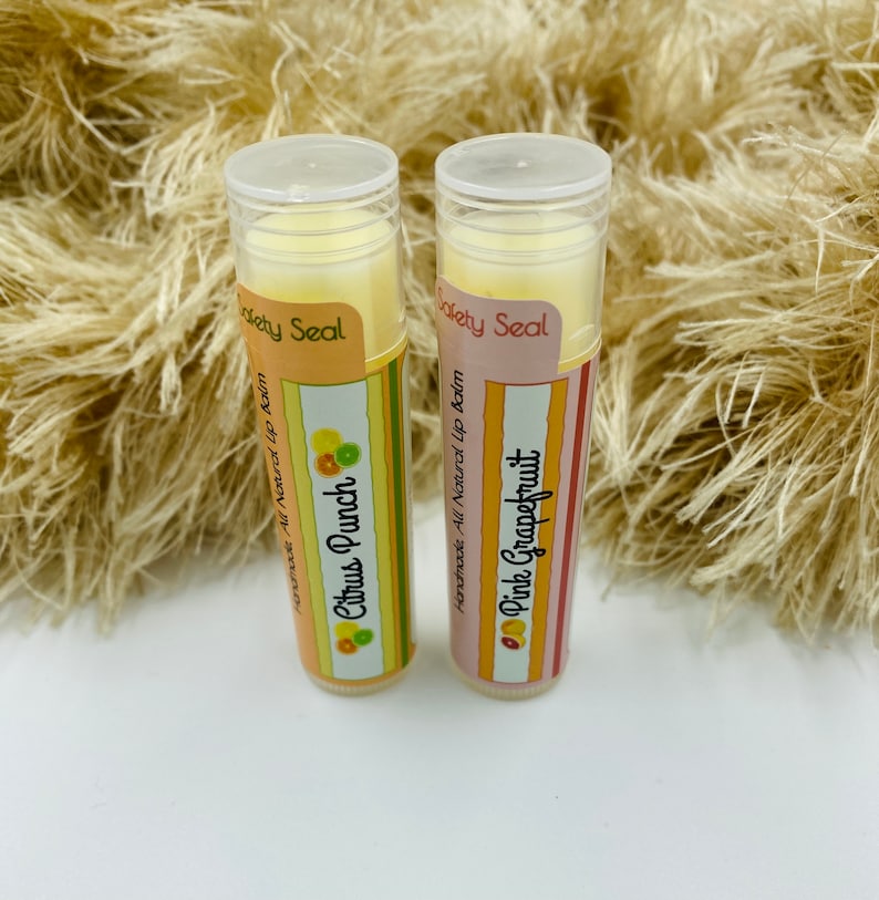 May include: Two tubes of lip balm with colorful labels. The lip balm on the left is labeled "Citrus Punch" and the lip balm on the right is labeled "Pink Grapefruit". Both tubes have a yellow and white striped label with the words "Handmade, All Natural Lip Balm" and "Safety Seal".