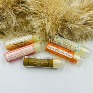 May include: Four different lip balm tubes with labels listing ingredients and contact information. The labels are in a gold and white color scheme with snowflakes and a handwritten font. The lip balms are all natural and handmade.