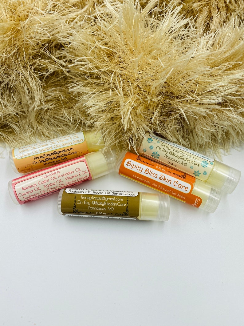 May include: Four different lip balm tubes with labels listing ingredients and contact information. The labels are in a gold and white color scheme with snowflakes and a handwritten font. The lip balms are all natural and handmade.