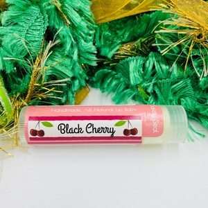 May include: A pink and white lip balm tube with a black cherry design and the text "Handmade. All Natural Lip Balm" and "Safety Seal".