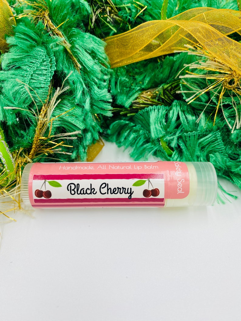 May include: A pink and white lip balm tube with a black cherry design and the text "Handmade. All Natural Lip Balm" and "Safety Seal".