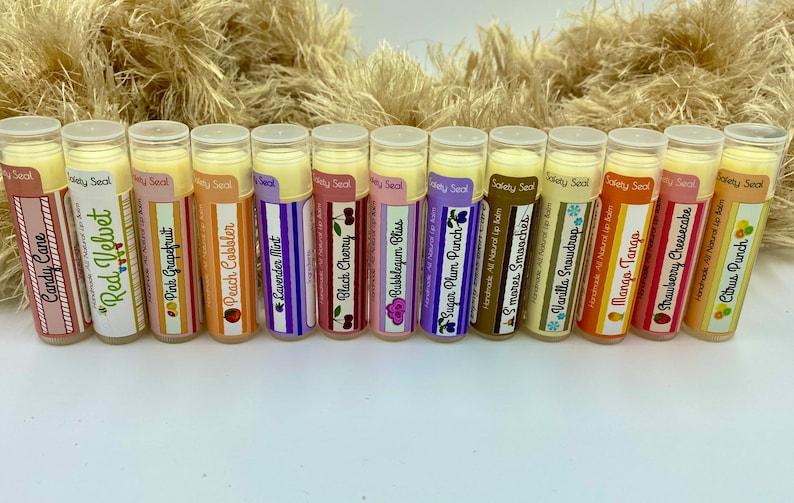 May include: A collection of 14 different flavored lip balms in clear tubes with colorful labels. The labels have names like "Candy Cane", "Red Velvet", "Pink Grapefruit", "Peach Cobbler", "Lavender Mint", "Black Cherry", "Bubblegum Bliss", "Sugar Plum Punch", "S'mores S'mores", "Vanilla Snowdrop", "Mango Tango", "Strawberry Cheesecake", and "Citrus Punch".