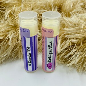 May include: Two lip balm tubes with purple and white labels. The labels say "Homemade All Natural Lip Balm" and "Safety Seal". The lip balm flavors are "Lavender Mint" and "Bubbleseum Bliss".