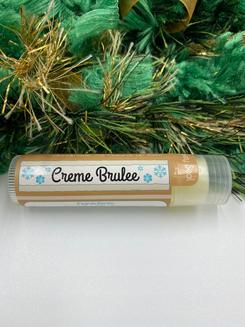 May include: A brown and white tube of lip balm with a snowflake design and the text "Creme Brulee" and "Handmade All Natural Lip Balm".