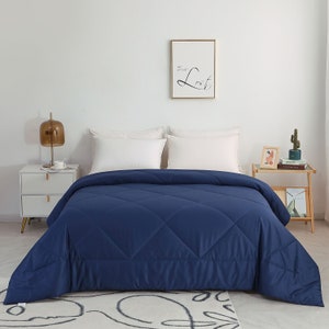 May include: A navy blue comforter with a diamond stitch pattern. The comforter is on a bed with white pillowcases and a white headboard. There is a framed print on the wall behind the bed.