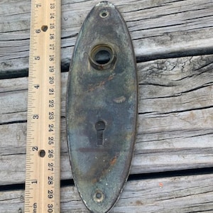 May include: An oval-shaped brass door plate with a keyhole and a round knob hole. The plate has a greenish patina and is slightly tarnished.