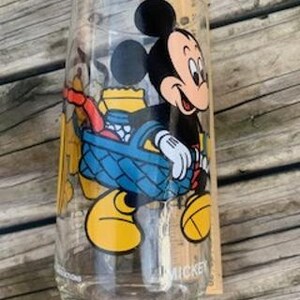 May include: A vintage glass drinking glass with a cartoon image of Mickey Mouse carrying a picnic basket. The glass is clear and has a Mickey Mouse logo at the bottom.