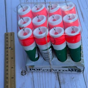 May include: A package of twelve cylindrical fishing floats. Each float has a red top, white middle section, and green bottom. The floats are packaged in a clear plastic container and are labeled "POP-R's FLOATS".