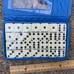 May include: A set of dominoes in a blue case. The dominoes are off-white with black dots. A wooden ruler is in the foreground. The case is open, revealing the dominoes arranged in rows.