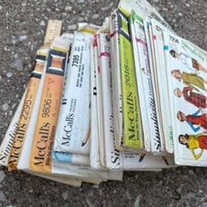 May include: A collection of assorted paper sewing patterns stacked together. The envelopes feature various brand names including Simplicity and McCall's, with visible model numbers like 2295, 9806, and 7395. Some envelopes display colorful illustrations of clothing designs.