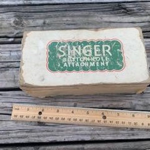May include: A vintage cardboard box for a Singer buttonhole attachment. The box is white with green lettering and a red border around the text. The text reads "Singer Buttonhole Attachment".