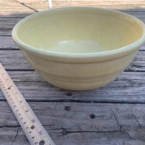 May include: A pale yellow ceramic mixing bowl with a smooth, rounded interior and a slightly flared rim. The bowl has a subtle, textured pattern on the exterior.