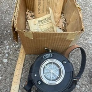 May include: A round black leather case with a silver-toned metal clock face featuring a protective grill, accompanied by a cardboard box containing paper instructions labeled TOP-OBEN and IMPORTANT, next to a wooden ruler for scale.