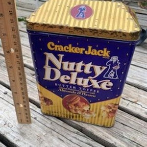 May include: A vintage Cracker Jack tin with a blue and gold design. The tin features the text "Nutty Deluxe" and "Butter Toffee" with the words "Almonds & Peanuts" below. The tin is sitting on a wooden surface.