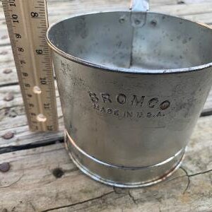 May include: A silver metal measuring cup with a handle. The cup has a black printed label that reads "Bromco Made in U.S.A."