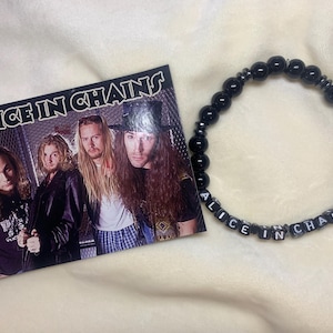May include: An Alice in Chains band photo and a black beaded bracelet. The bracelet features the band's name spelled out in white letter beads. The photo shows the band members with long hair.