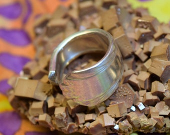 Könnte beinhalten: Ein silberner Ring mit einem einzigartigen Design liegt auf einem Bett aus braunen, unregelmäßig geformten Kristallen. Die Kristalle sind in einer Gruppe angeordnet und bilden einen strukturierten und organischen Hintergrund.