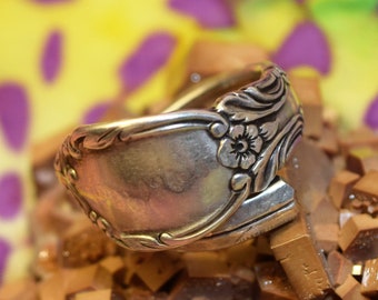 Könnte beinhalten: Ein silberner Löffelring mit floralem Design. Der Ring liegt auf einem Haufen kleiner, brauner, rechteckiger Stücke.