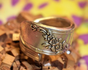 Könnte beinhalten: Ein silberner Ring aus einer wiederverwendeten Löffel mit einem floralen Design. Der Ring liegt auf einem Bett aus braunen Kristallen.