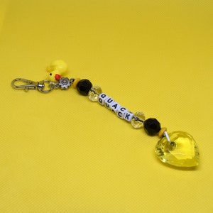 以下が含まれることがあります： 赤いクチバシと黒い目をした黄色いゴム製のオモチャのカモのチャームが、銀色のキーホルダーに取り付けられています。キーホルダーには、黒と透明なビーズと、白い文字ビーズが「QUACK」と綴られています。キーホルダーの端には、黄色のクリスタル製のハートのチャームが付いています。