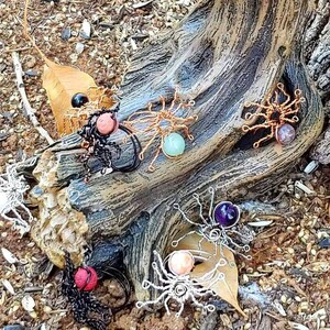 Puede incluir: Una colección de anillos de araña de alambre hechos a mano con piedras preciosas de varios colores. Los anillos se muestran en un tronco de árbol con un fondo natural.