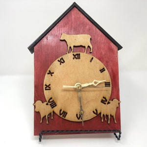 May include: A red wooden clock shaped like a house with a cow and sheep cutouts. The clock face is light brown with black Roman numeral markings.