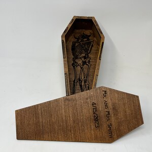 May include: A wooden coffin-shaped box with a skeleton couple inside. The lid of the box is open and the lid has the text "MR. AND MRS. SMITH 4/15/2023" engraved on it.