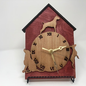 May include: A wooden clock shaped like a doghouse with a red stained finish. The clock face is made of light wood with black Roman numeral hour markers and black hands. The clock is surrounded by three dog silhouettes.