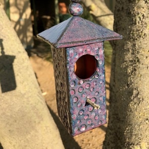May include: A purple and blue ceramic birdhouse with a round opening and a textured surface. The birdhouse is hanging from a string with a series of purple and blue beads.