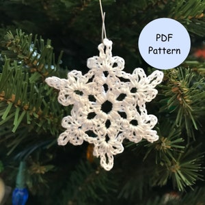 以下が含まれることがあります： 緑のクリスマスツリーに吊るされた白いクロシェの雪の結晶のオーナメント。PDFパターン