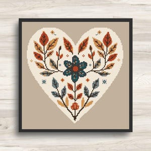 Può includere: Un motivo a punto croce incorniciato a forma di cuore, caratterizzato da un fiore centrale blu scuro con centro arancione, circondato da disposizioni simmetriche di foglie arancioni, marroni e blu e piccoli accenti floreali geometrici su sfondo crema.