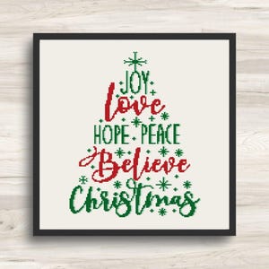 May include: A framed Christmas-themed cross-stitch design. The artwork features a Christmas tree shape composed of the words "JOY, LOVE, HOPE, PEACE, BELIEVE, CHRISTMAS" in red and green. The background is off-white.