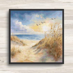 Puede incluir: Una obra de arte en punto de cruz enmarcada que representa una escena de playa serena. Un camino arenoso conduce al mar, flanqueado por dunas y hierbas. El cielo está lleno de nubes, gaviotas y el brillo del sol. El marco es negro.