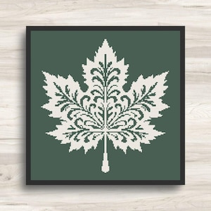 May include: A square framed cross-stitch pattern featuring a white maple leaf with intricate, symmetrical scrollwork details set against a solid dark green background. The design is contained within a thin black border, creating a clean visual.