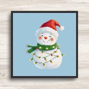 May include: A cross-stitch image of a smiling snowman wearing a red Santa hat, green scarf, and a string of yellow lights. The snowman has rosy cheeks and a carrot nose. The background is a light blue color.