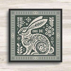 May include: A framed cross-stitch design featuring a detailed rabbit illustration in cream and dark green. The rabbit is surrounded by floral and geometric patterns, set against a dark green background with a cream border.
