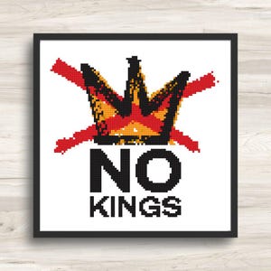 May include: A framed cross-stitch art piece with a white background. The design features a pixelated crown in black, orange, and red, crossed out with red lines. Below the crown, the words "NO KINGS" are displayed in black block letters.