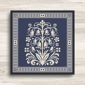 Op de afbeelding: Een ingelijst kruissteekpatroon met een gestileerd bloemmotief in crème tegen een marineblauwe achtergrond. Het ontwerp omvat klokvormige bloemen en decoratieve randen, alles binnen een zwart frame. Gepresenteerd op een licht houten oppervlak.