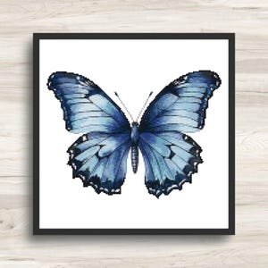 Op de afbeelding: Een vierkant ingelijst wandkunstwerk met een gedetailleerd kruissteekpatroon van een vlinder. De vlinder toont symmetrische vleugels in verschillende tinten blauw, variërend van licht hemelsblauw tot diep marineblauw, met zwarte omlijningen en details.