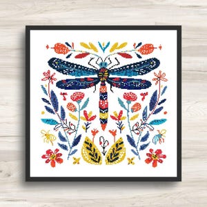 Op de afbeelding: Een ingelijst kruissteekpatroon met een libel met blauwe vleugels en een gestreept lichaam. De libel is omgeven door kleurrijke bloemen, bladeren en insecten in rood, geel en blauw. Het kunstwerk staat tegen een witte achtergrond.