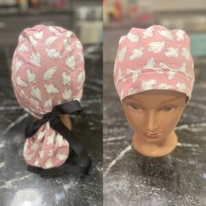May include: Pink surgical scrub hat with a white ghost pattern. The hat has a black tie that can be tied at the back of the head.