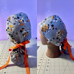 May include: A gray and orange surgical scrub hat with a Halloween Mickey Mouse print. The hat has a tie at the back and is made of a soft, comfortable fabric.