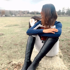 May include: A woman wearing a blue long-sleeved shirt with white text, white riding trousers, and black riding boots sits on a bale of hay. The shirt has the text "INJEE" and "CIC" on one sleeve and "NIEG" on the other sleeve.