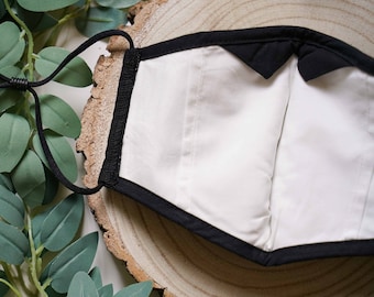 May include: Black face mask with white lining and adjustable ear loops. The mask is lying on a wooden surface.