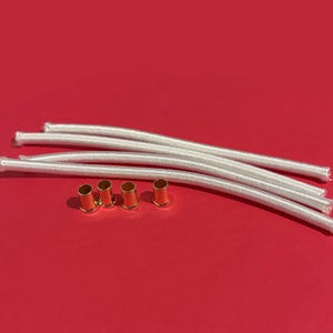 May include: A collection of white elastic cords and gold-colored metal eyelets against a red background. The cords are arranged in a curved pattern, and the eyelets are lined up below them. The image is well-lit and the items are in focus.