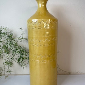 May include: A tall, cylindrical mustard yellow ceramic vase with a narrow neck and a flared rim. The exterior surface features a repeating embossed pattern consisting of horizontal bands of diamonds, small rectangles, and dots in a grid layout.