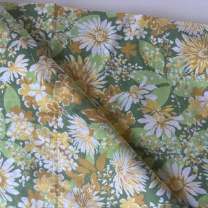 May include: A folded fabric featuring a dense floral pattern with white, mustard yellow, and light green daisy-like flowers and leaves. The textile displays a repeating botanical print across the entire surface, showing a soft texture and drape.