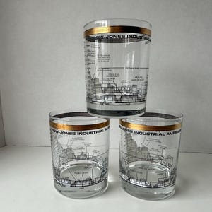 May include: Three clear glass tumblers featuring a gold rim and black printed graphics depicting the Dow-Jones Industrial Average. The design includes historical timeline markers, stock market charts, and text labels such as Apollo 11 Man on Moon.